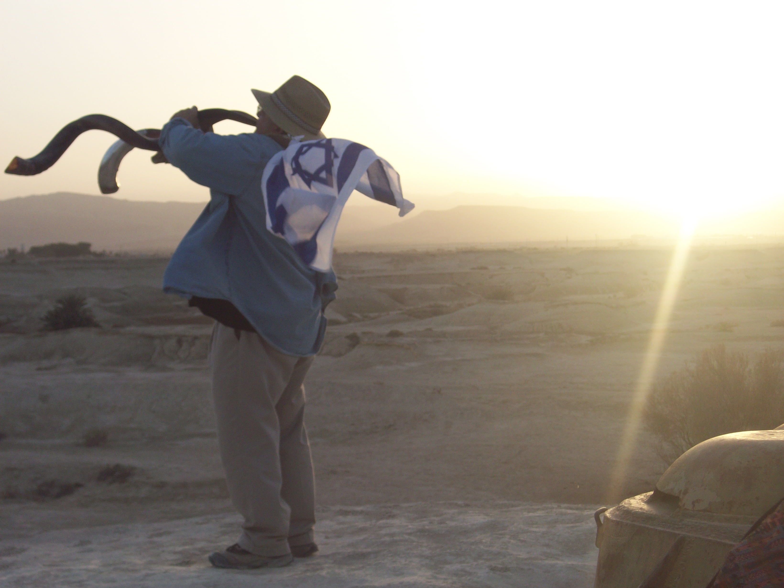 Robert Weinger sounding the shofar over the desert at sunset, Israeli flag draped over his shoulders, Beit Hogla landscape stretching to the mountains