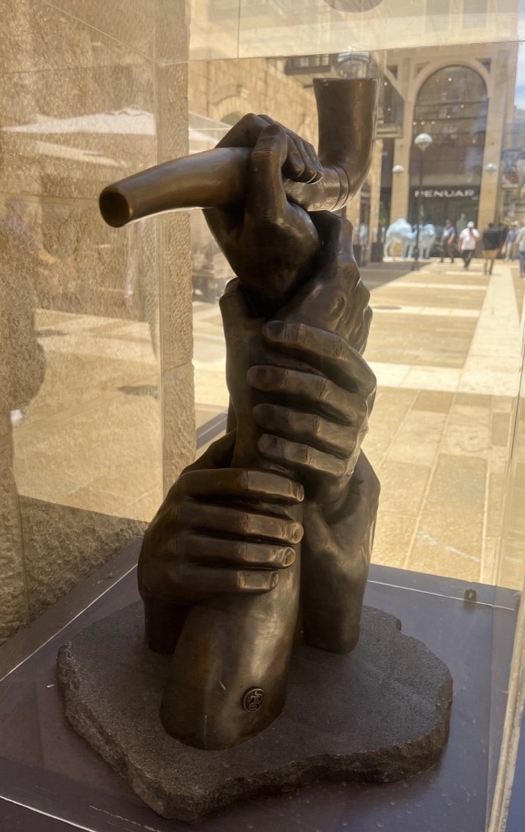 The Great Shofar bronze sculpture installed in its glass case on Mamilla Avenue, Jerusalem — intertwined hands lifting a shofar, the Mamilla promenade visible beyond